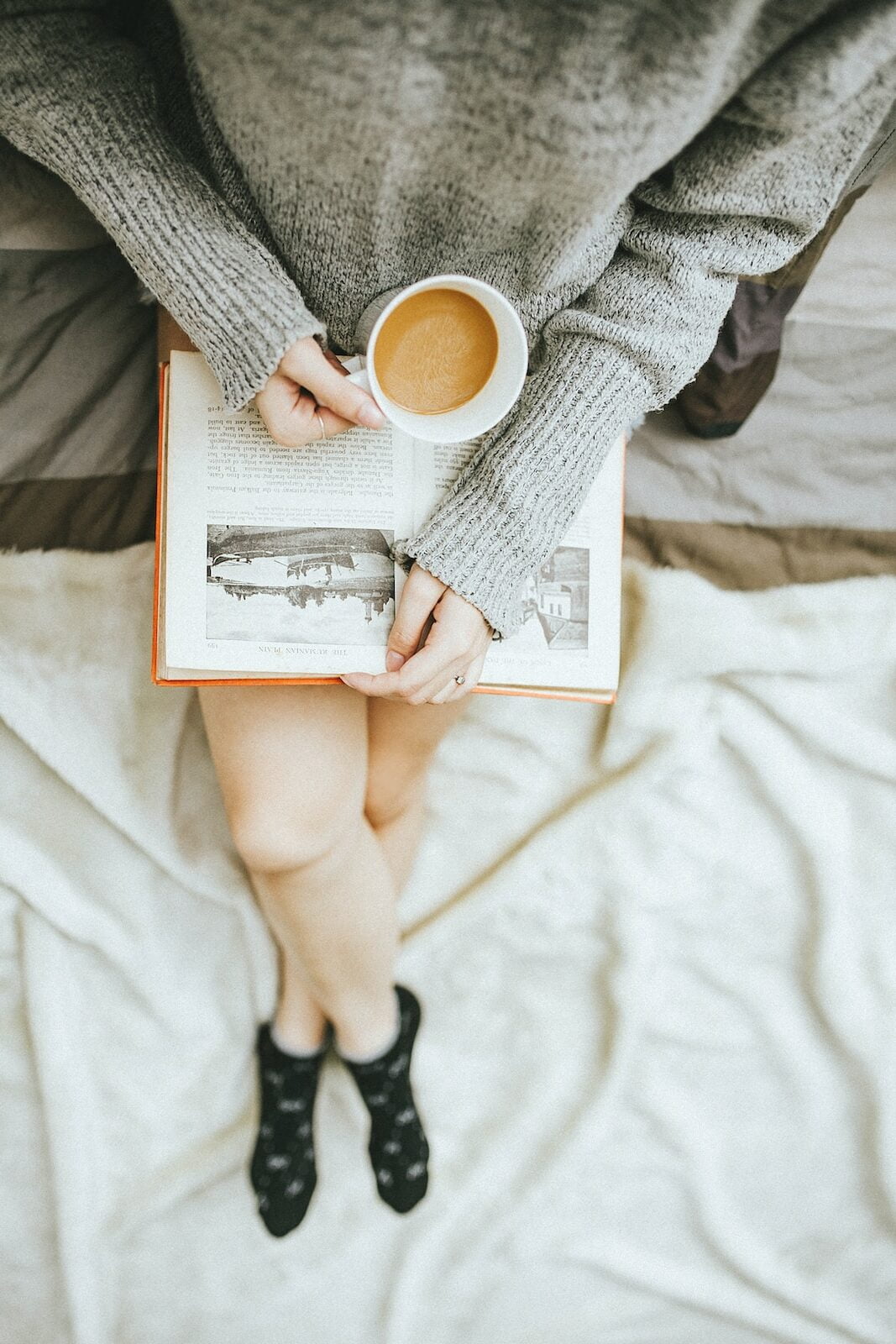 woman holding a cup of coffee at right hand and reading book on her lap while holding it open with her left hand in a well-lit room